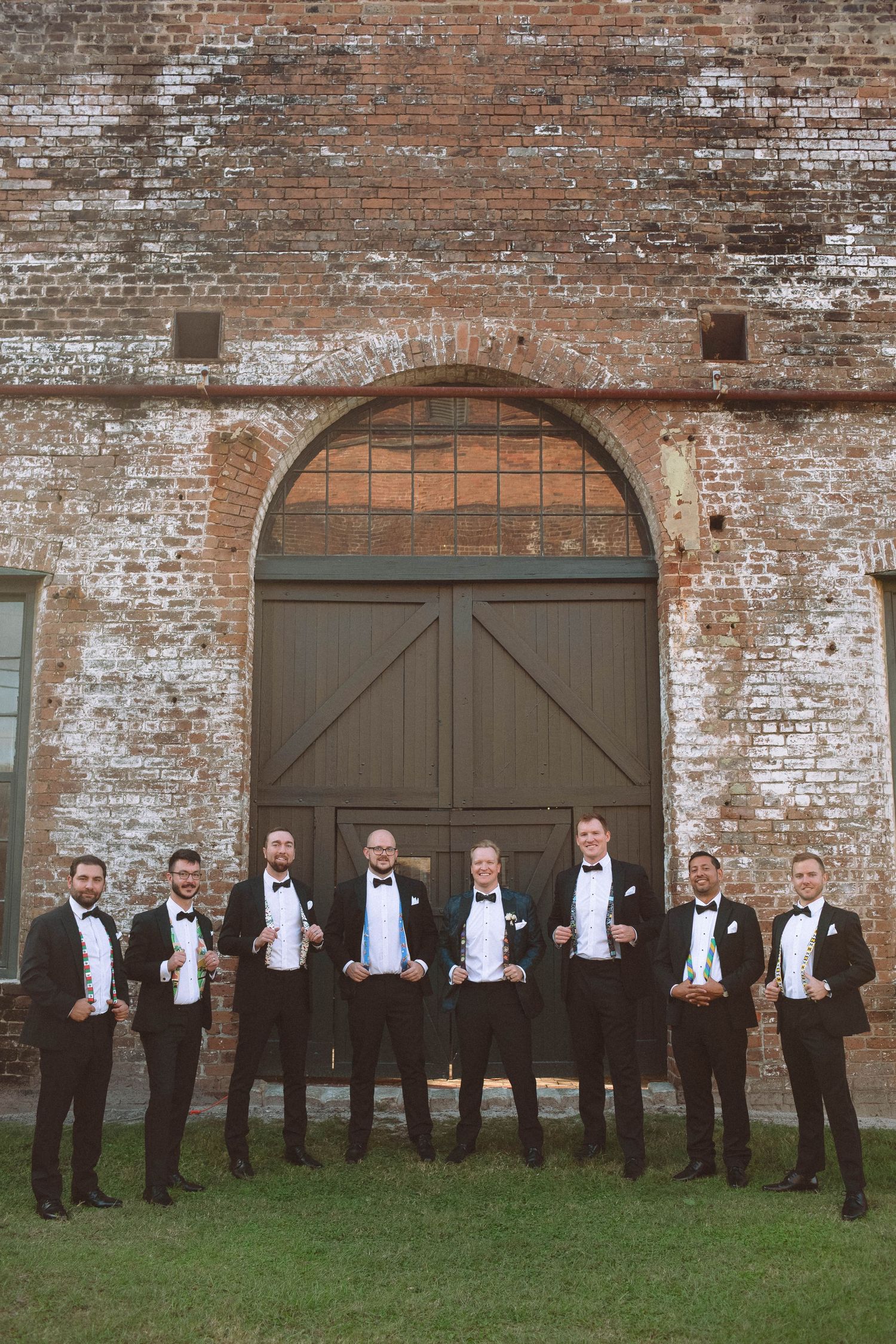 Groomsmen in black tuxedos holding drinks outside a rustic brick building with arched doors.