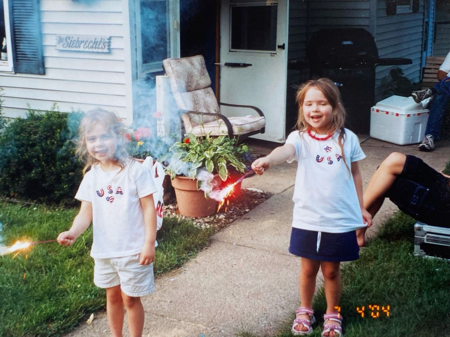Two young girls in matching USA shirts holding sparklers on a driveway during Fourth of July.