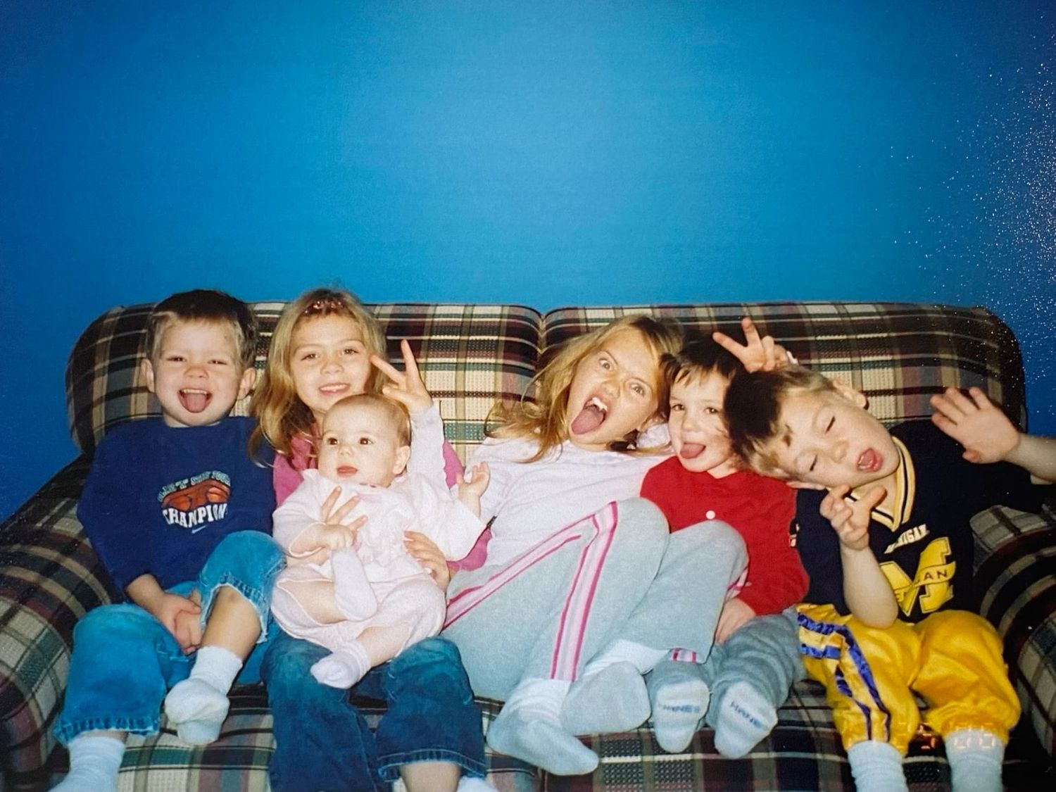 Six children pose playfully on a plaid couch in front of a bright blue wall.