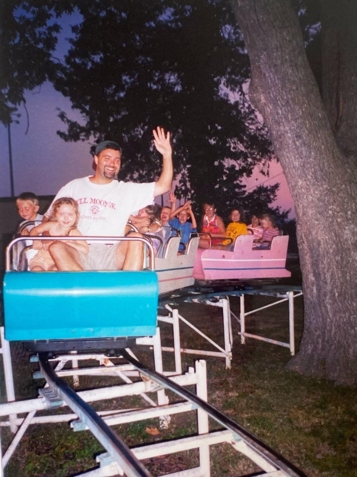 A man waving cheerfully while riding a small roller coaster with children at an amusement park.