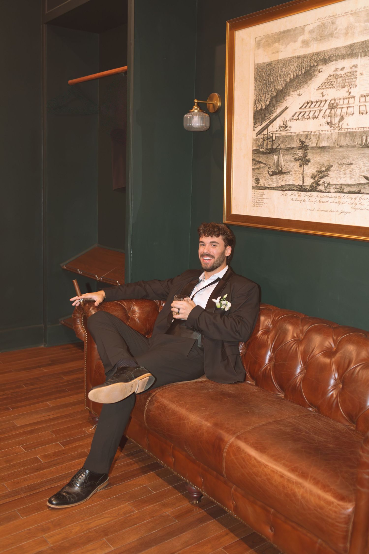 Smiling groom in dark suit relaxing on leather Chesterfield sofa with boutonniere and vintage wall art.