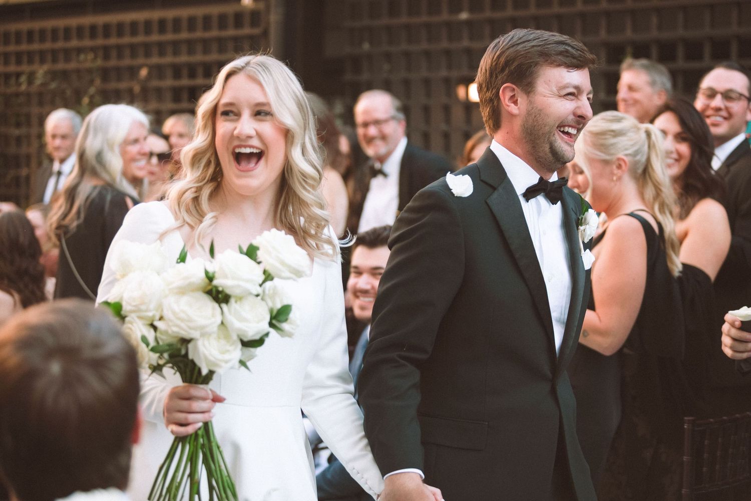 Joyful bride laughing holding white rose bouquet with groom in tuxedo after ceremony.