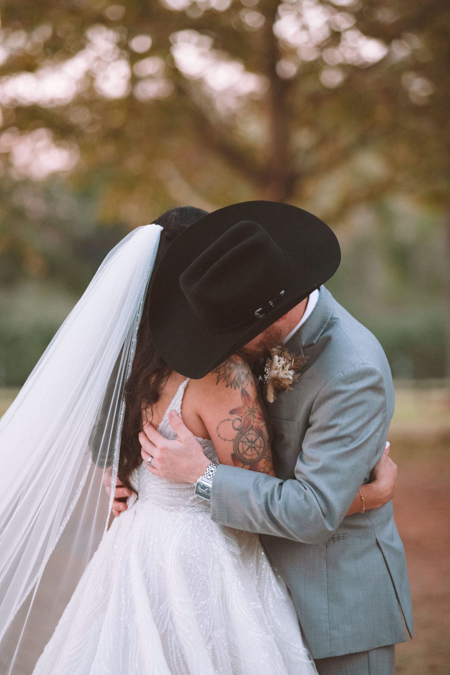 Bride and groom embrace in intimate first kiss, groom wearing black cowboy hat outdoors.
