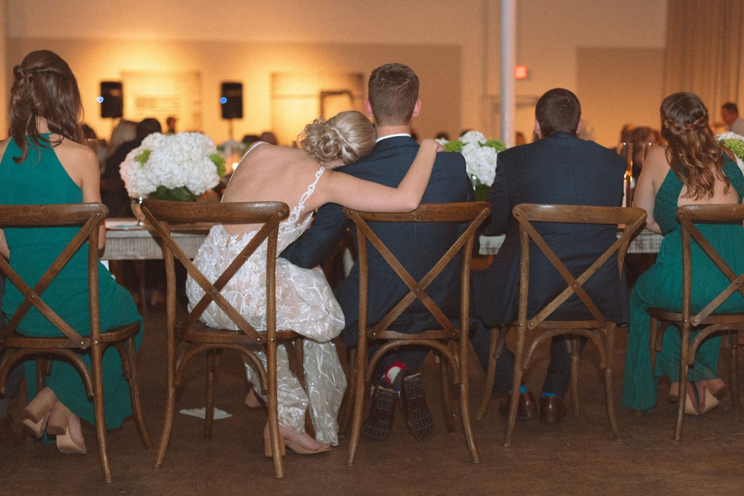 Bride and groom sit closely together at sweetheart table surrounded by wedding party in teal.