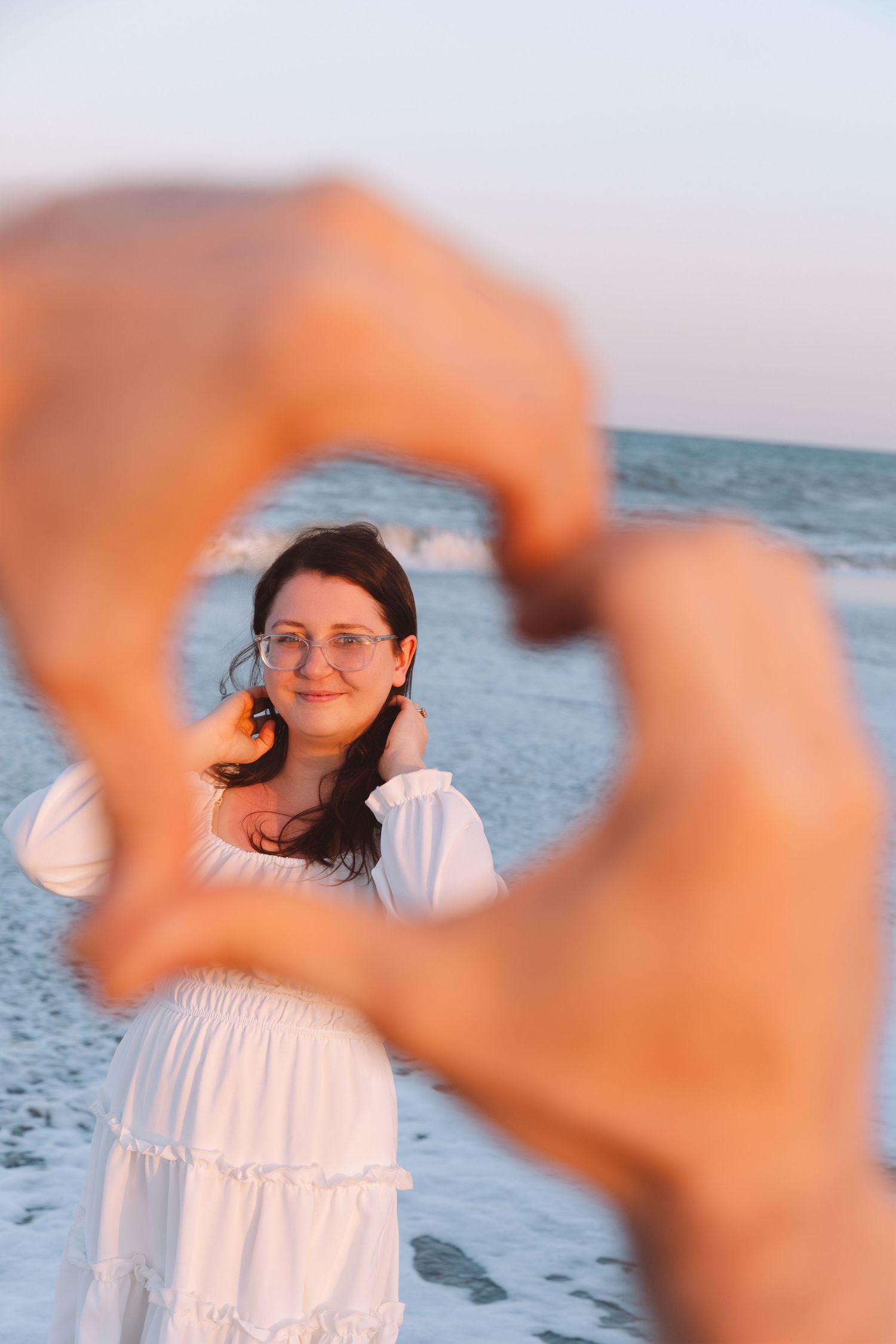 Woman in white dress framed by heart-shaped hands at sunset beach, smiling through glasses with ocean waves behind her.