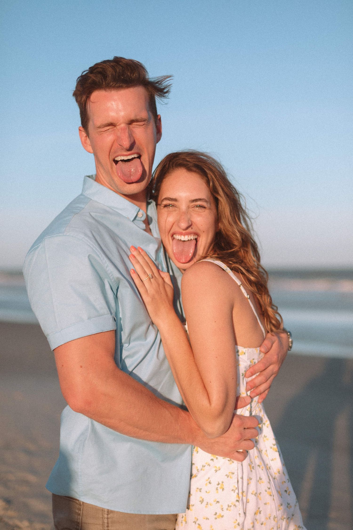 Happy couple laughing and sticking out tongues while embracing on a sunny beach at golden hour.