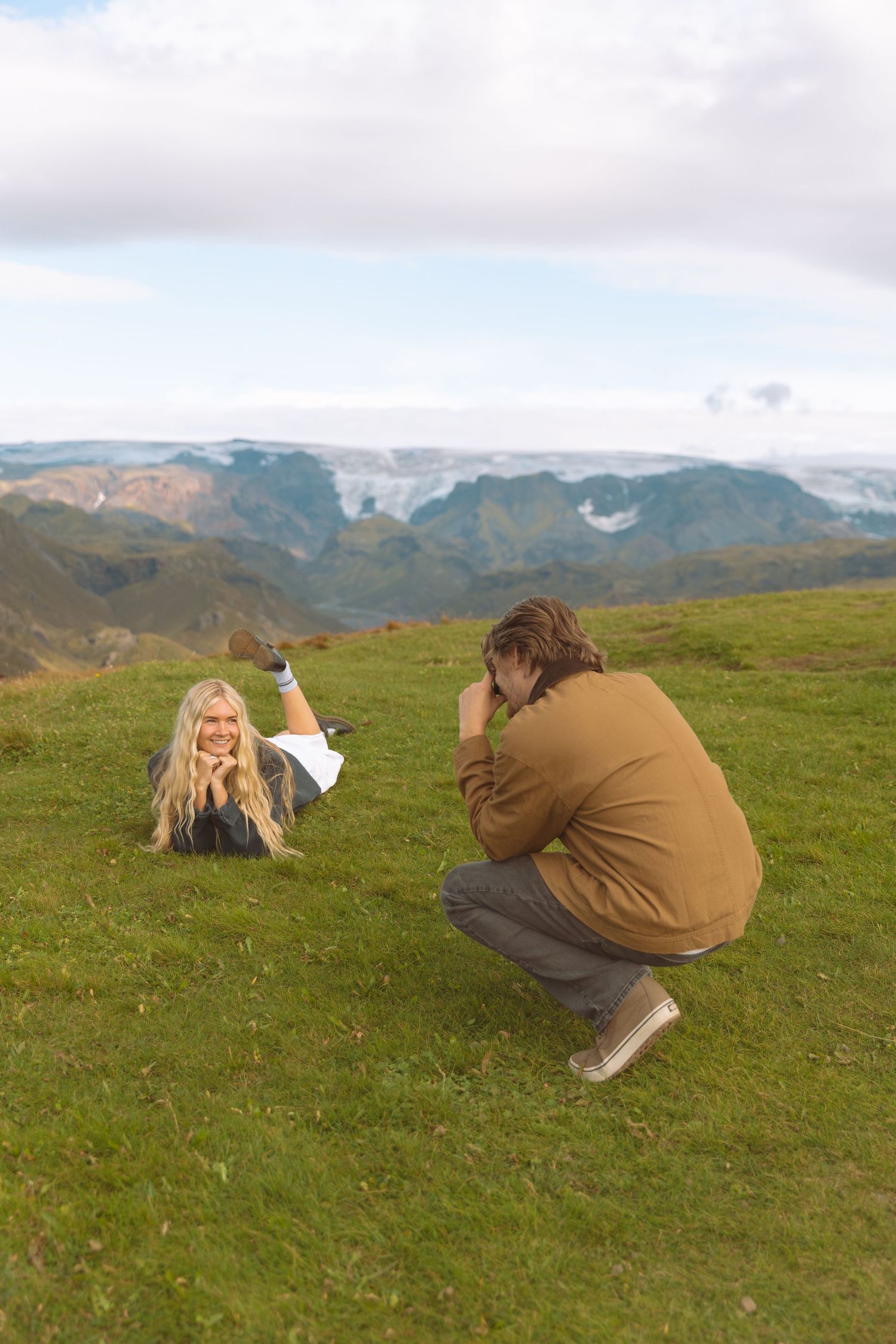 Photographer crouching to capture smiling blonde woman lying on mountain hillside.