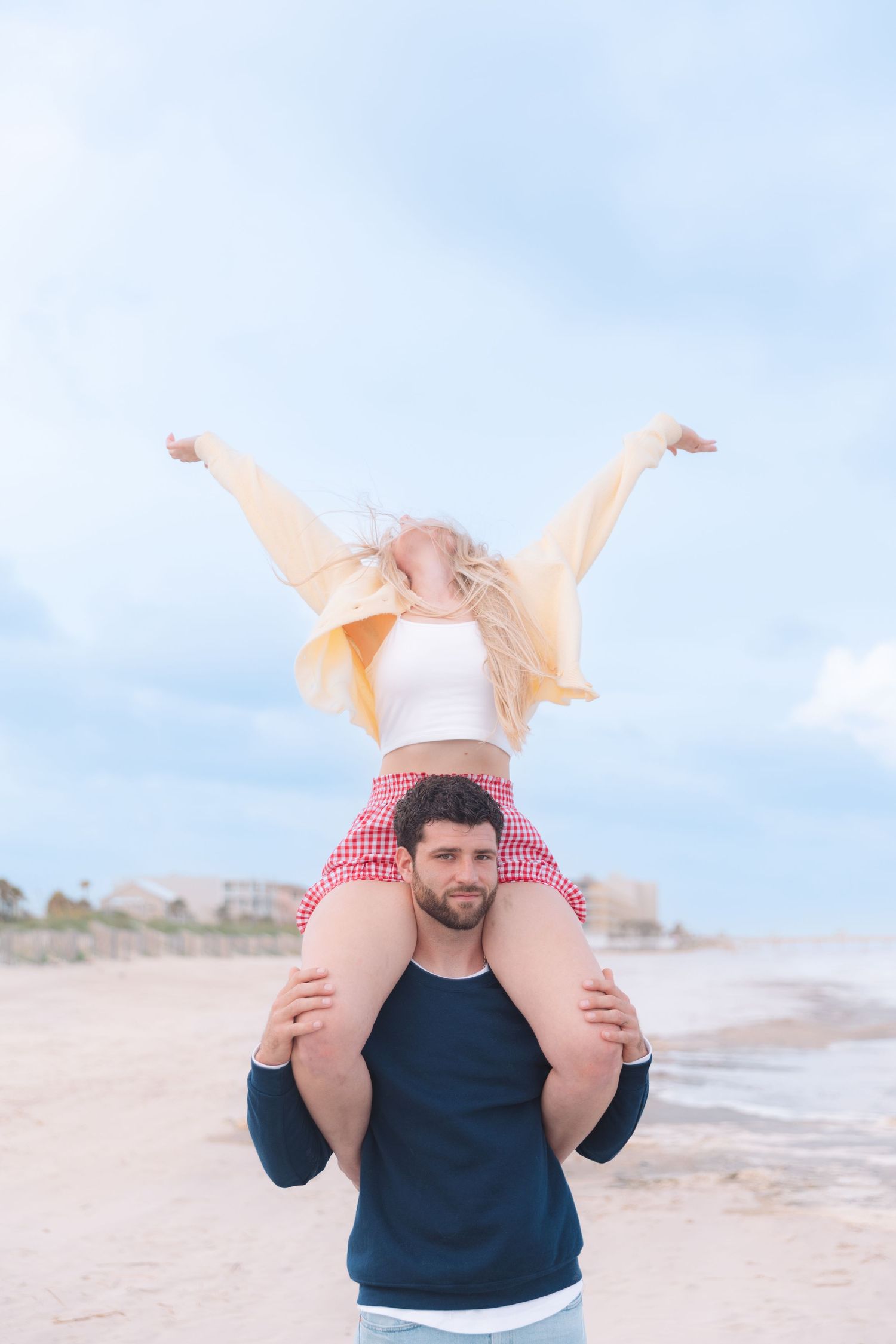 Man carrying woman on his shoulders at the beach, both smiling and enjoying a fun sunny day by the ocean.