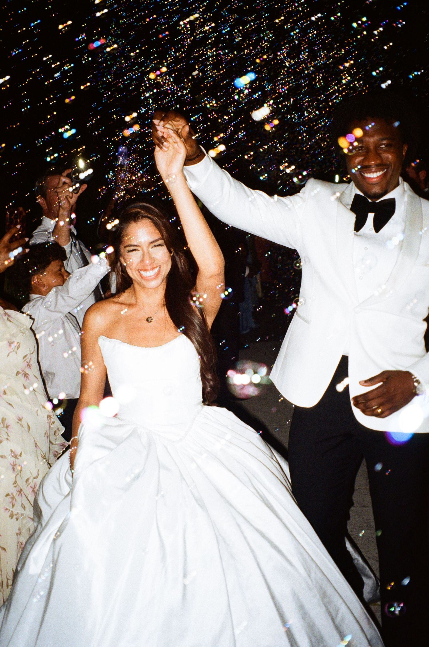 Joyful newlyweds celebrate with raised hands during festive wedding reception with bokeh lights and bubbles.