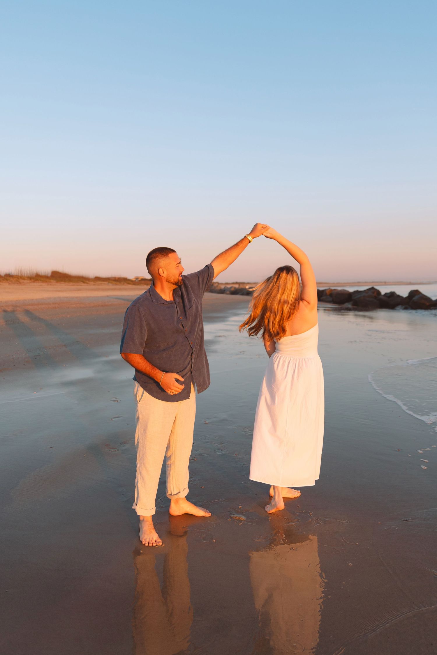 Couple dancing barefoot on a sandy beach at golden hour sunset, man spinning woman in white dress by ocean waves.