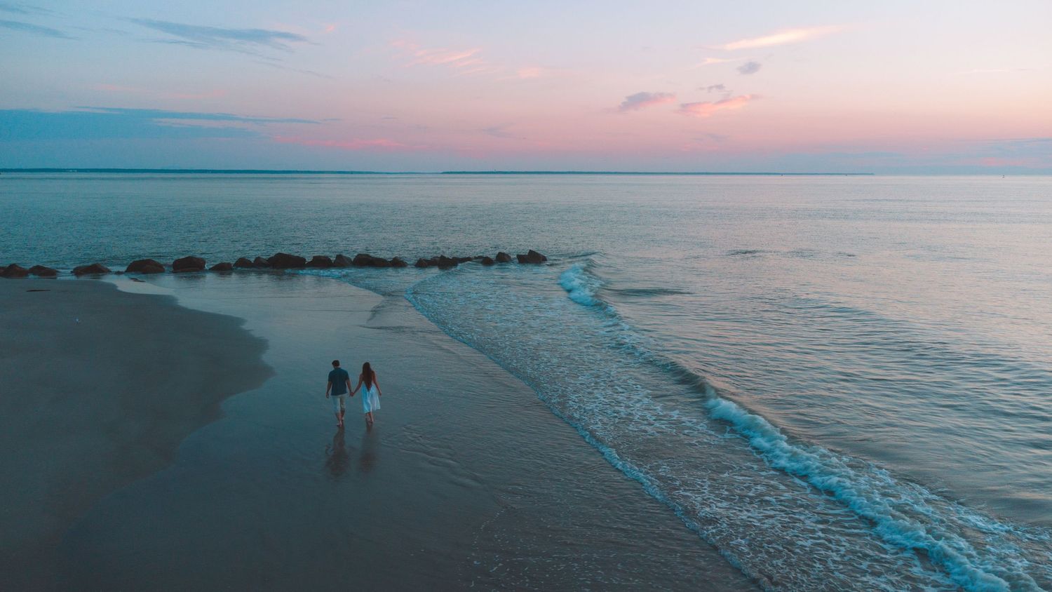 Couple walking hand in hand on a sandy beach at sunset with pastel pink and blue sky reflecting on calm ocean waters.