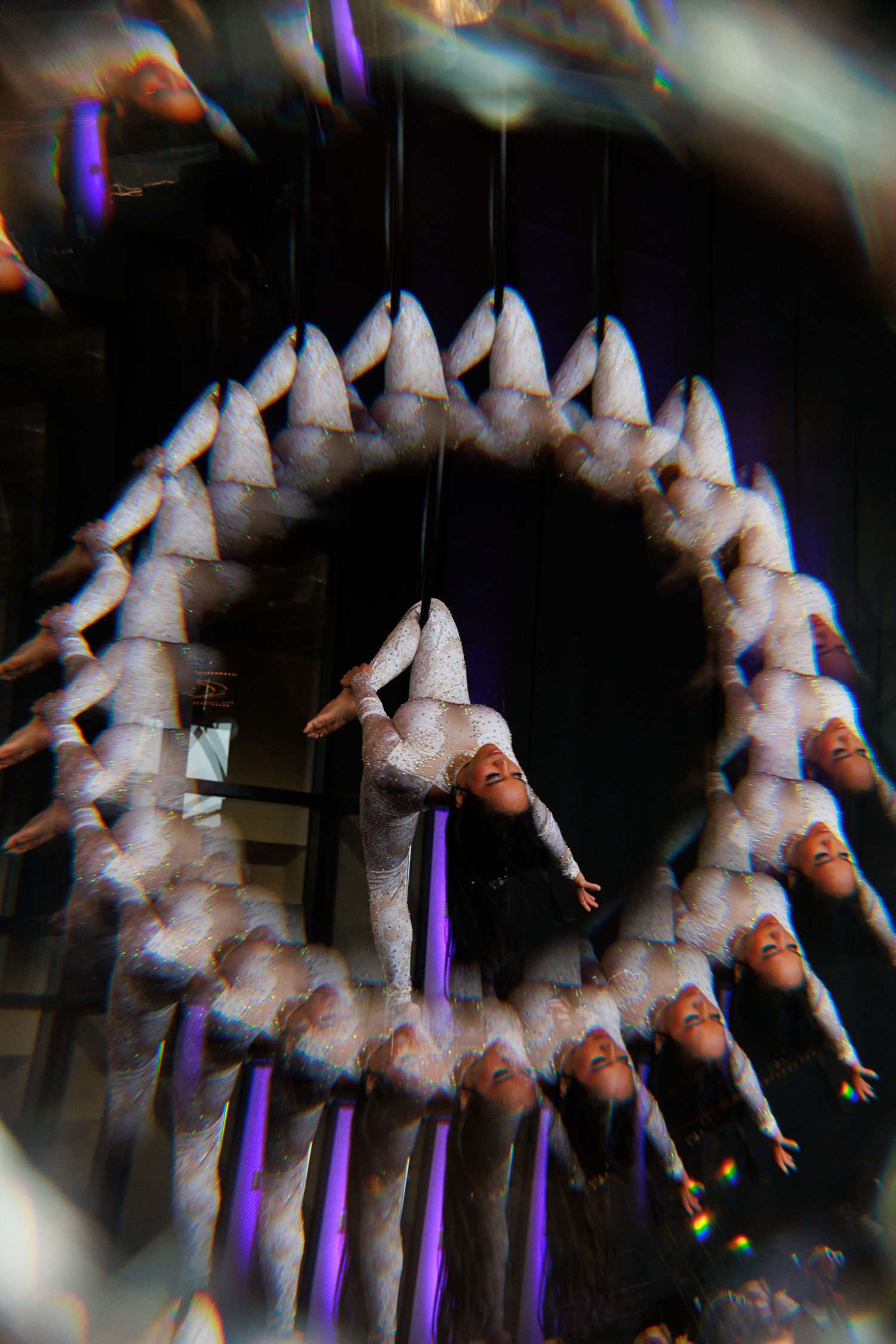 Aerial acrobat performing in white costume with multiple exposure effect creating circular pattern against dark background.