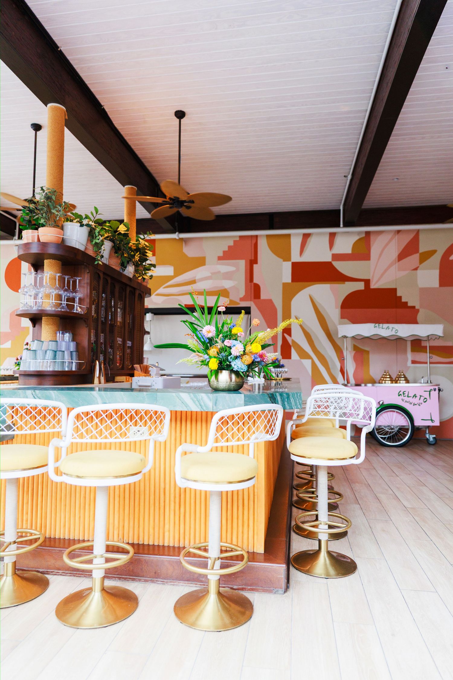 Colorful retro bar with gold swivel stools, geometric mural, marble countertop, and tropical floral arrangement.