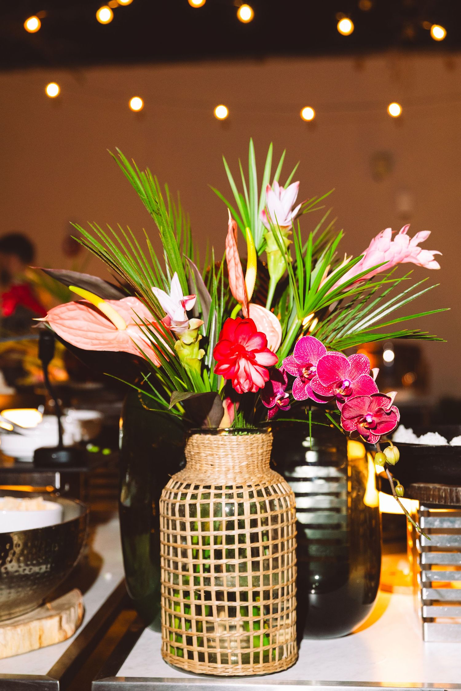 Tropical floral centerpiece with orchids and palm leaves in a woven bamboo vase on a table.