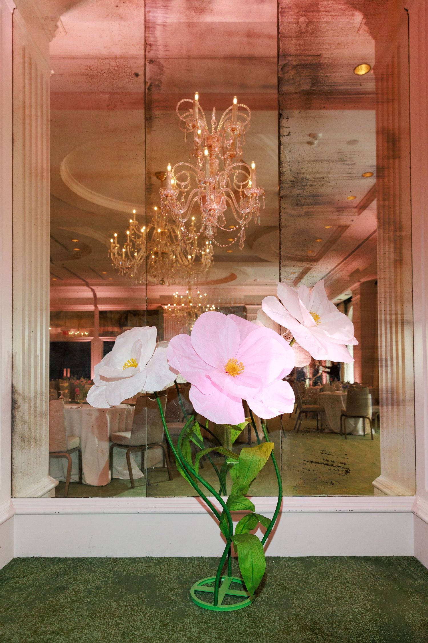 Large decorative pink and white flowers in a green vase outside an elegant ballroom with chandelier.