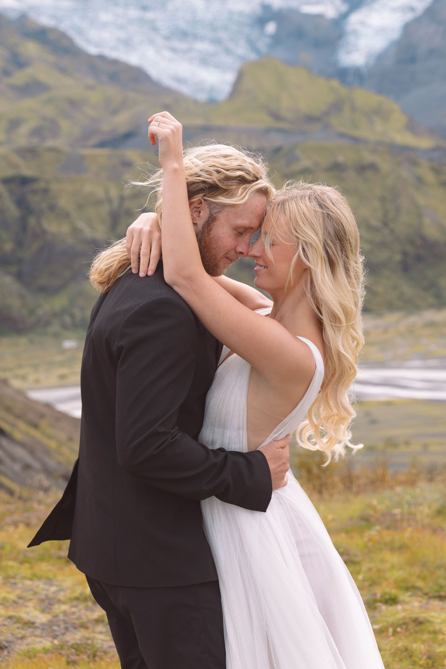 Bride and groom embrace romantically with foreheads touching against a stunning mountain glacier backdrop.