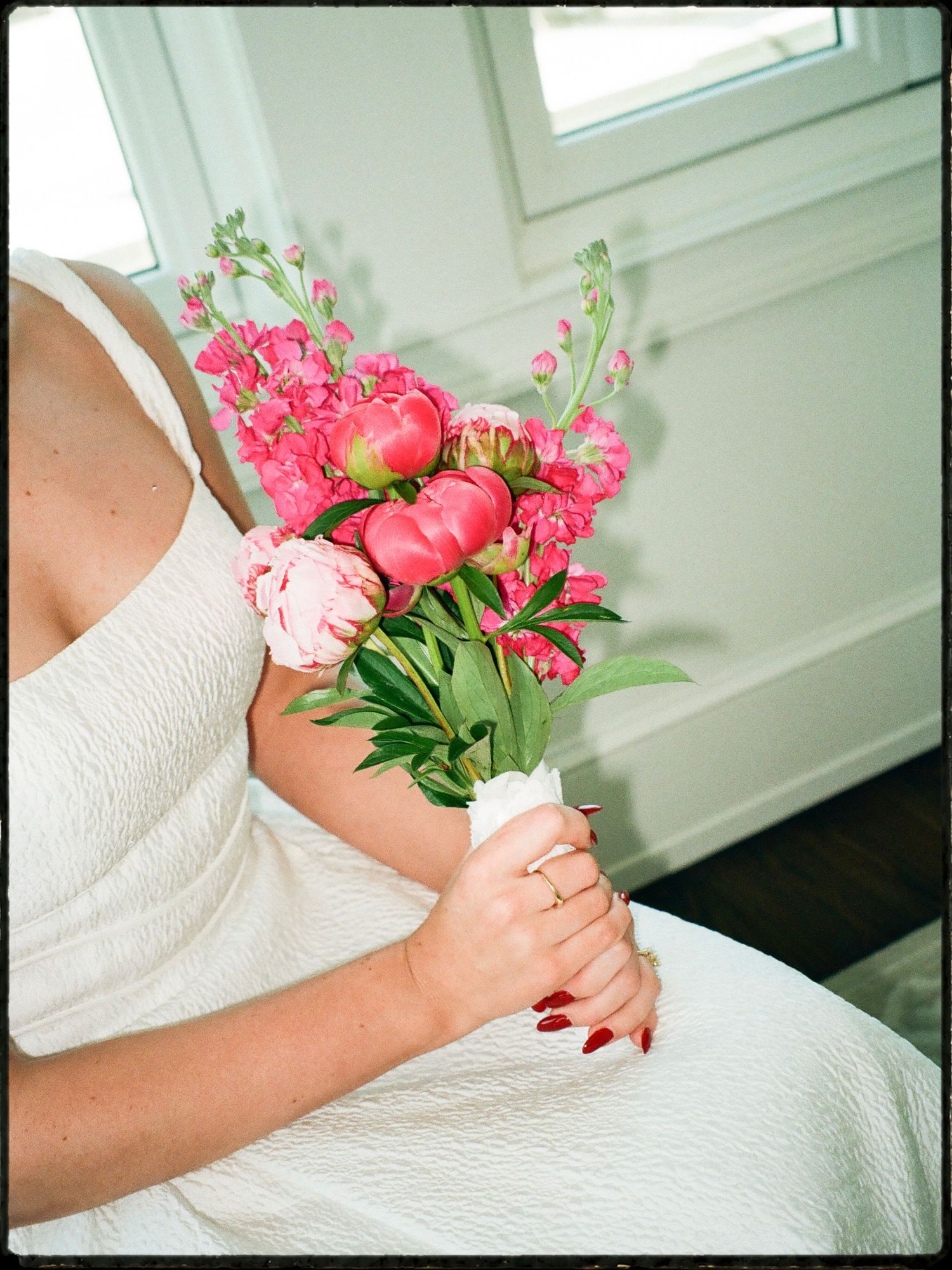 Bride in white dress holding a vibrant pink peony and flower bouquet wrapped in white ribbon.