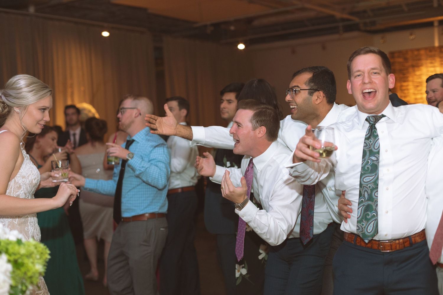 Wedding guests laughing and dancing with drinks at a lively indoor reception celebration.