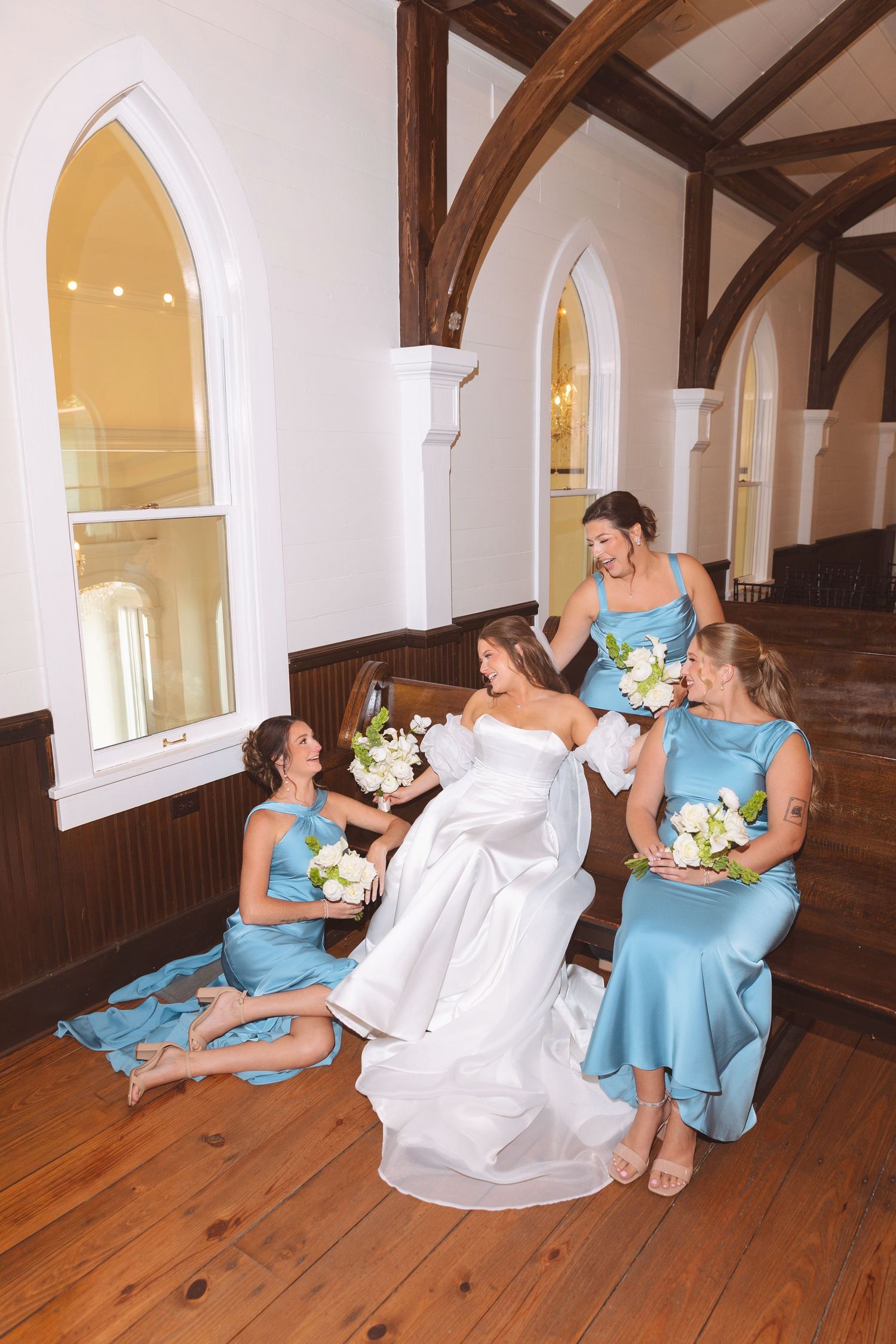 Bride in white gown laughing with three bridesmaids in blue dresses inside historic church.