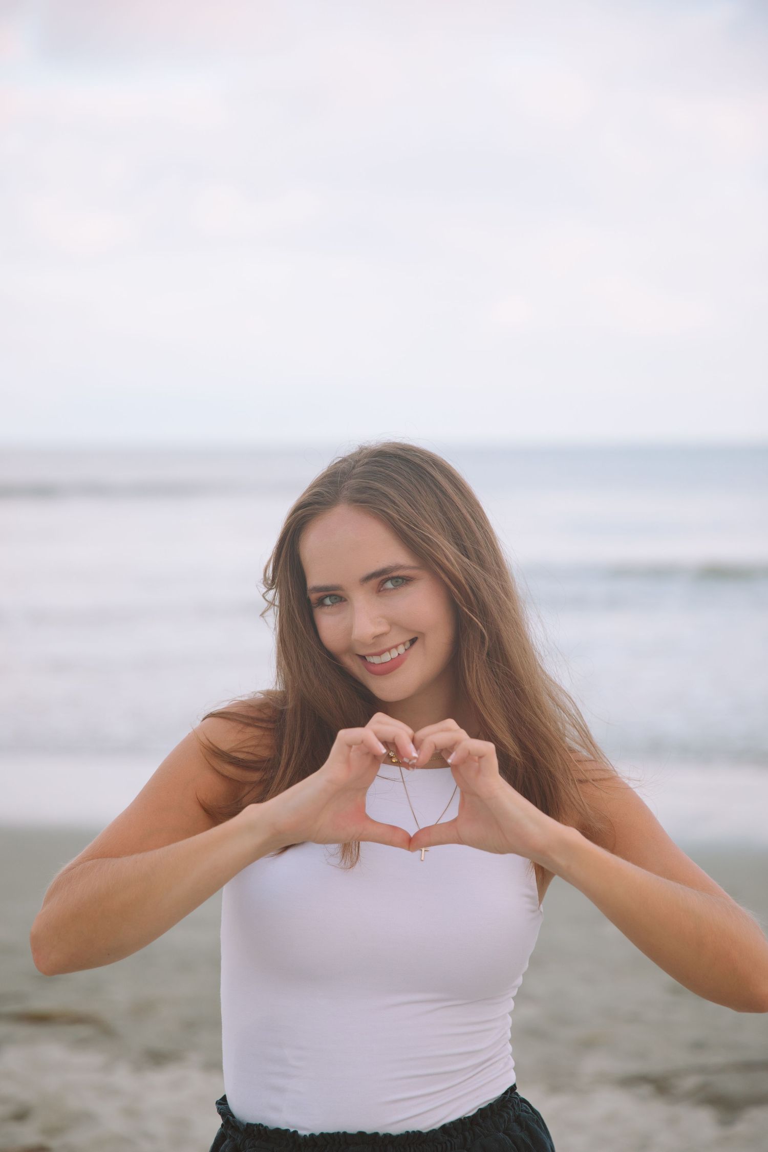 Young woman smiling on beach making heart shape with hands, wearing white tank top with ocean in background.