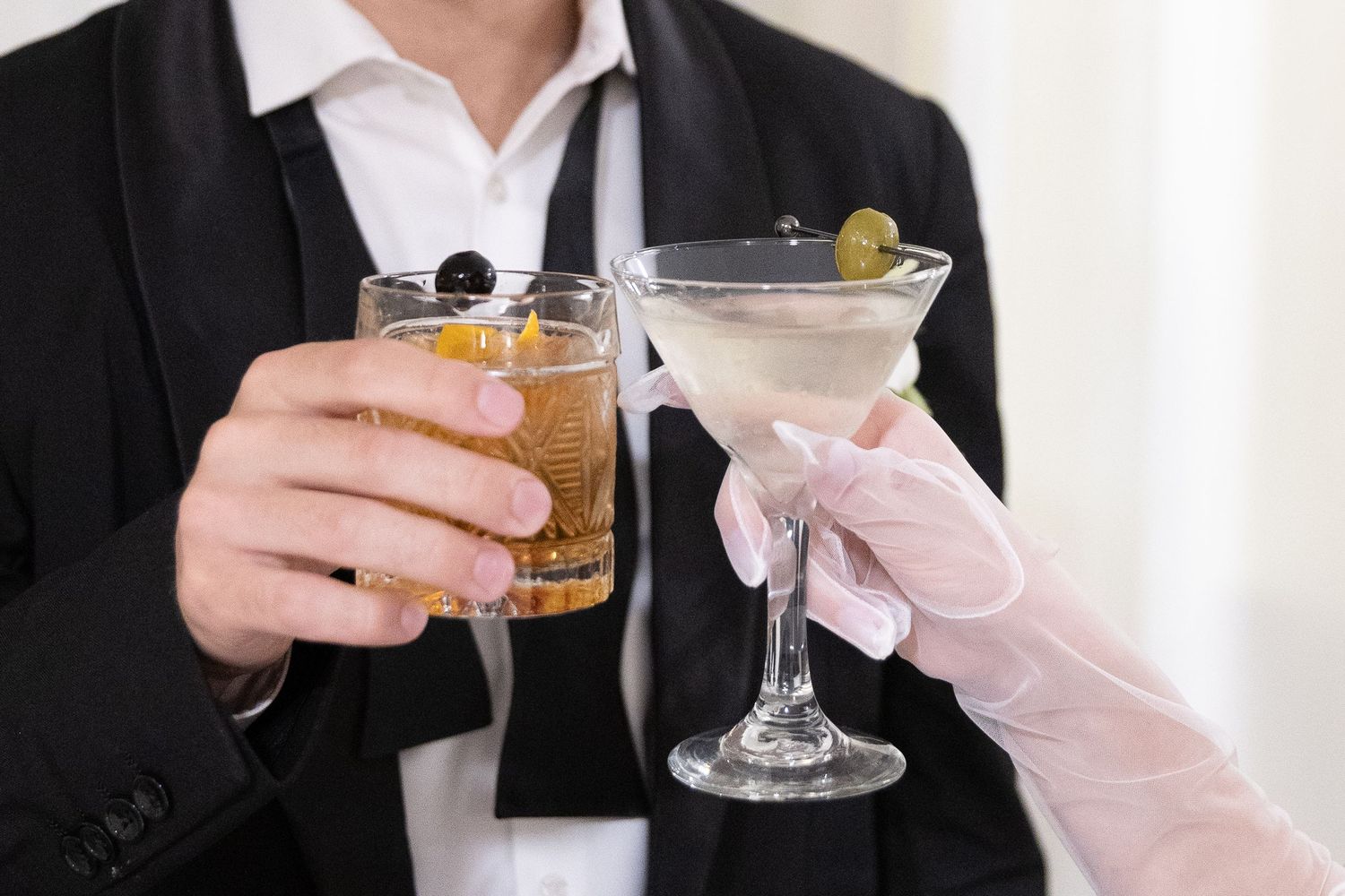 Two people clinking cocktail glasses, one an old fashioned and one a martini, at a formal event.