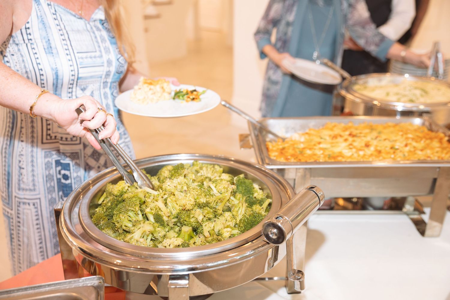 Woman serving herself broccoli rice dish from a round silver chafing dish at a buffet.