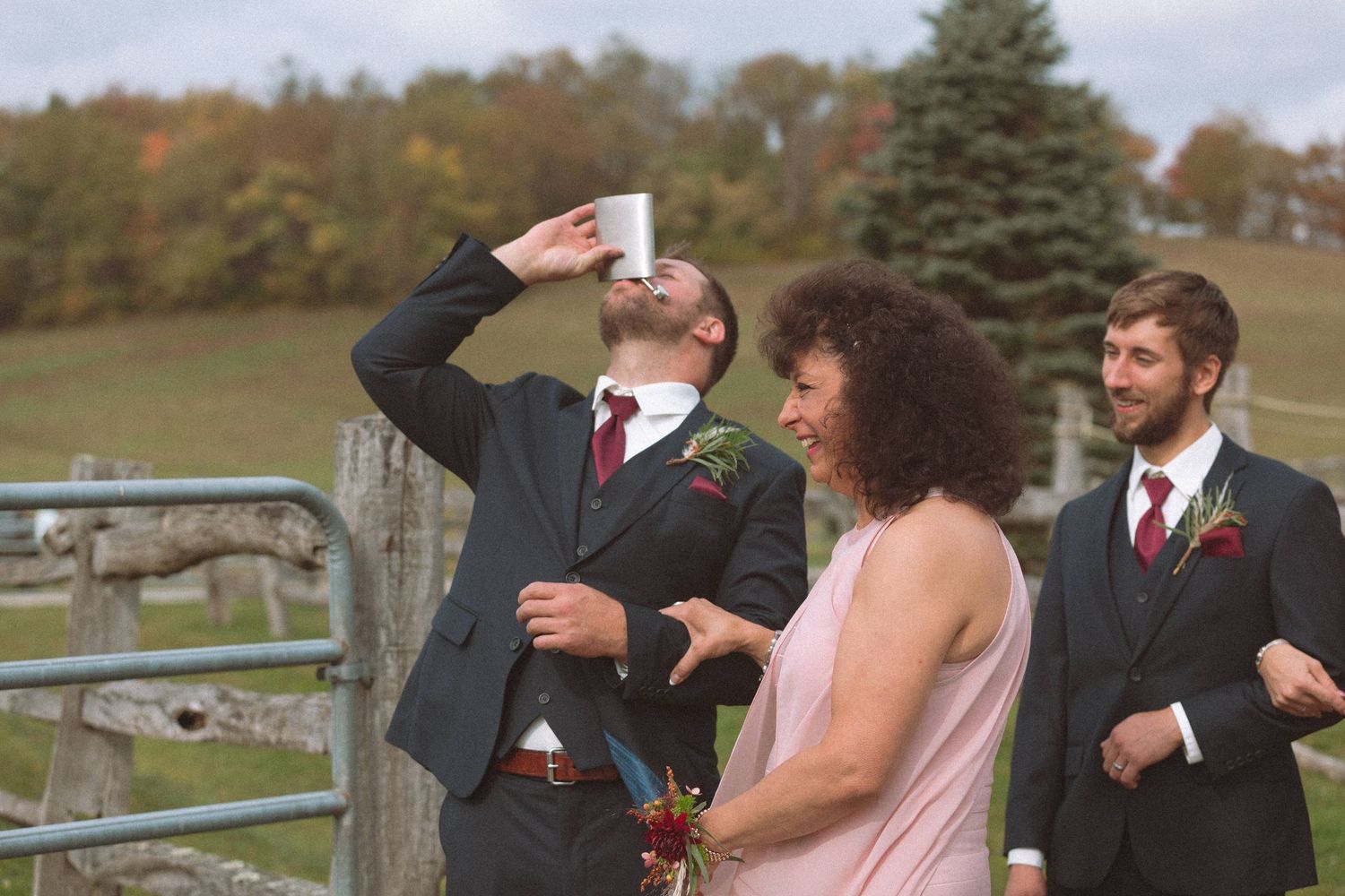 Groomsman taking a shot before wedding ceremony while others watch and laugh outdoors.