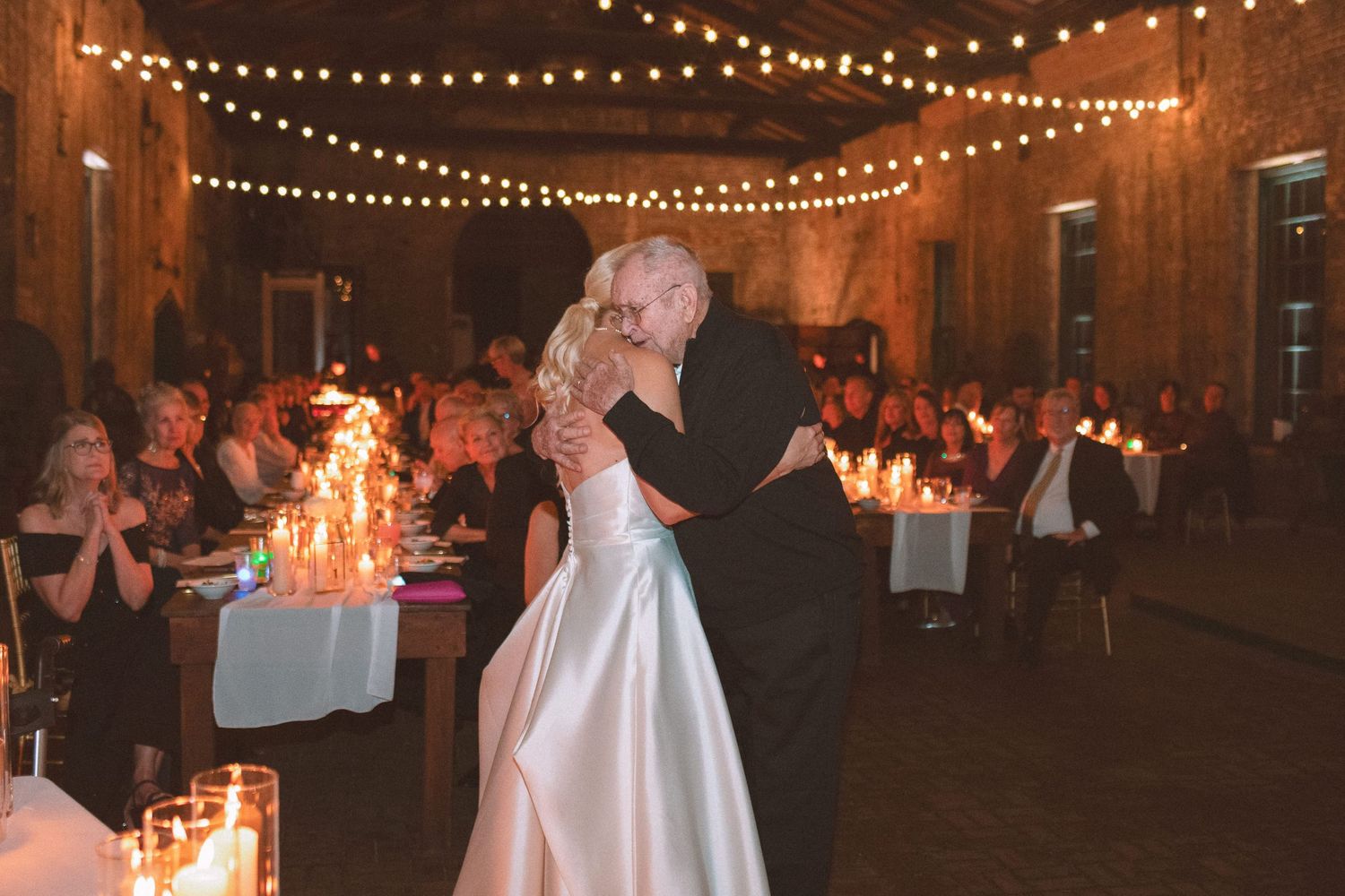 Bride and groom sharing a romantic first dance kiss under string lights in a candlelit barn venue.