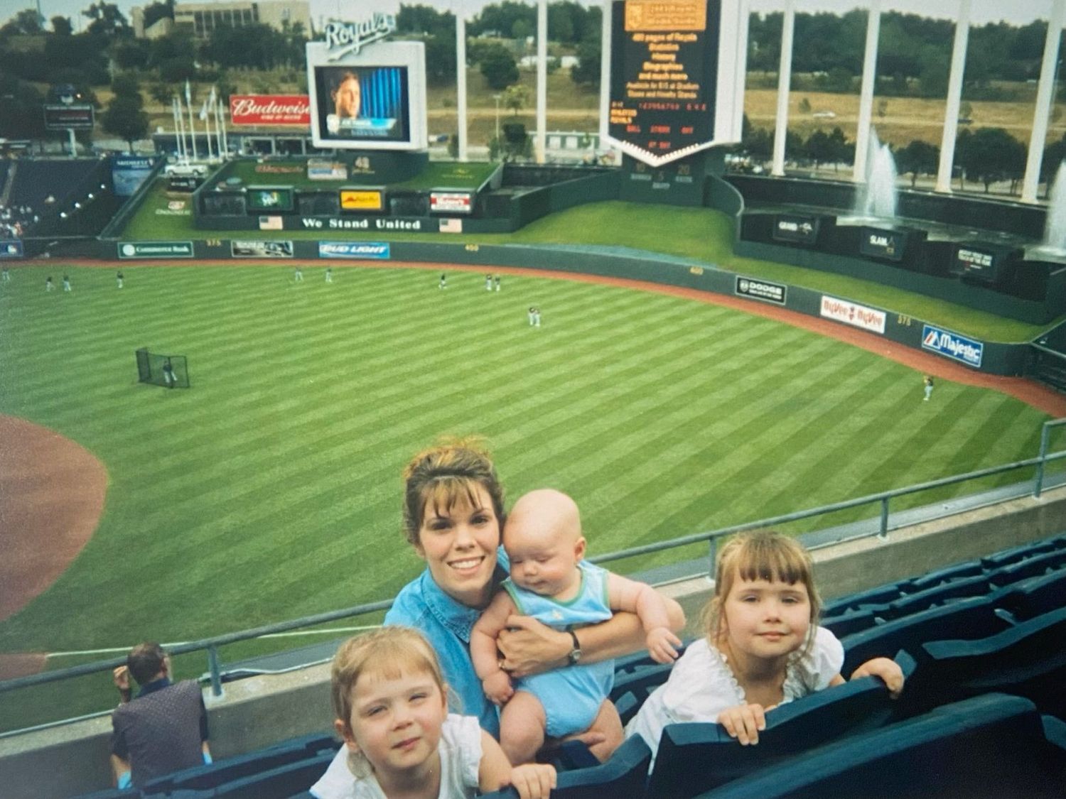 Mother holding baby with two children posing at a baseball stadium overlooking the field.