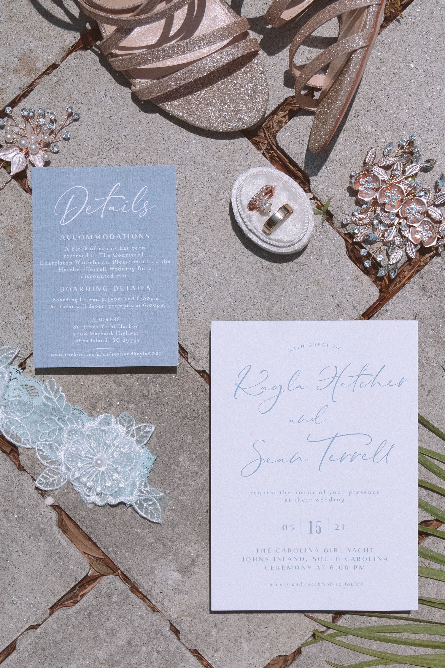 Wedding flat lay featuring dusty blue invitation suite, rose gold rings, lace garter, and floral hair accessories.