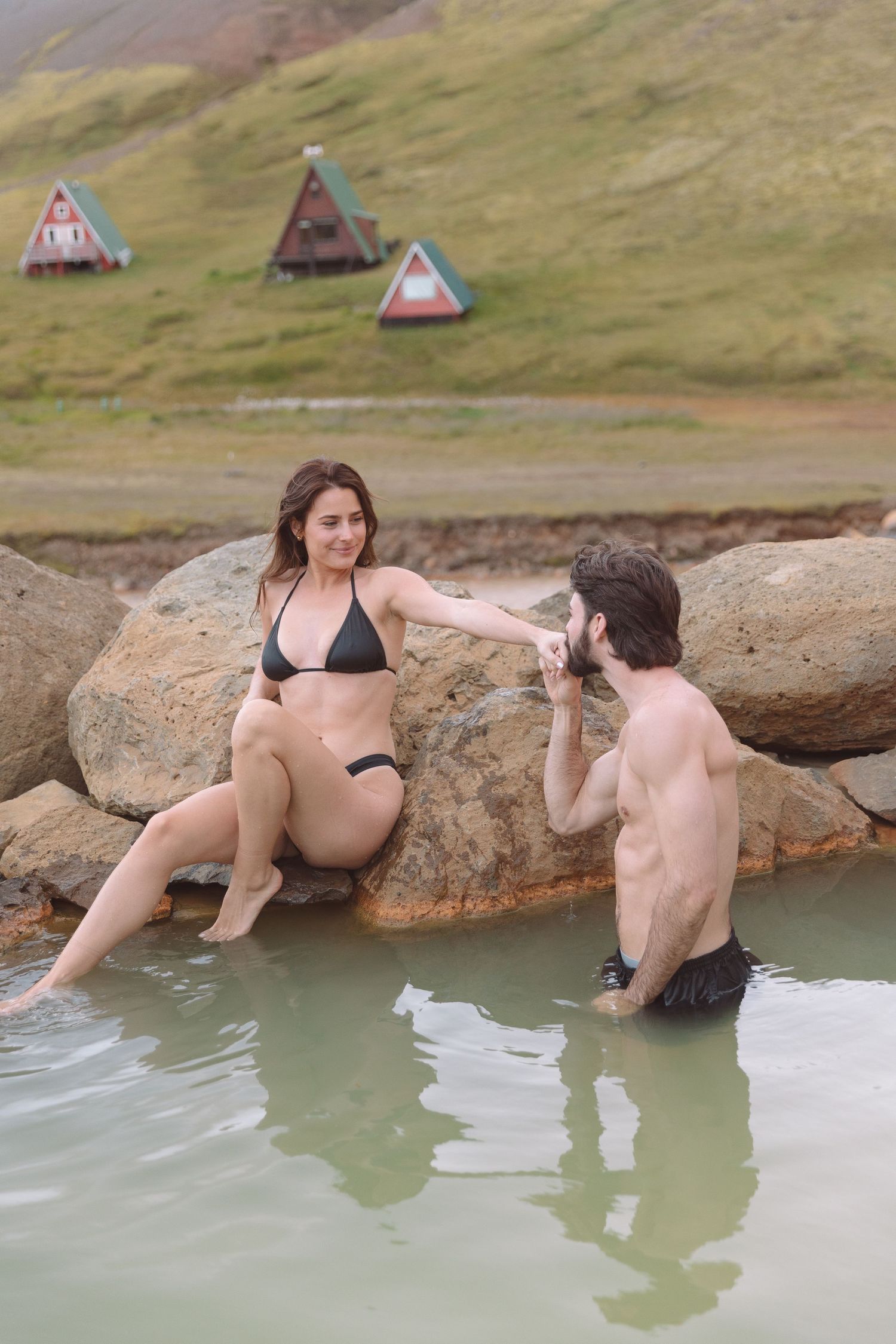 Man playfully splashing water toward woman sitting on rocks at a natural geothermal pool in Iceland.