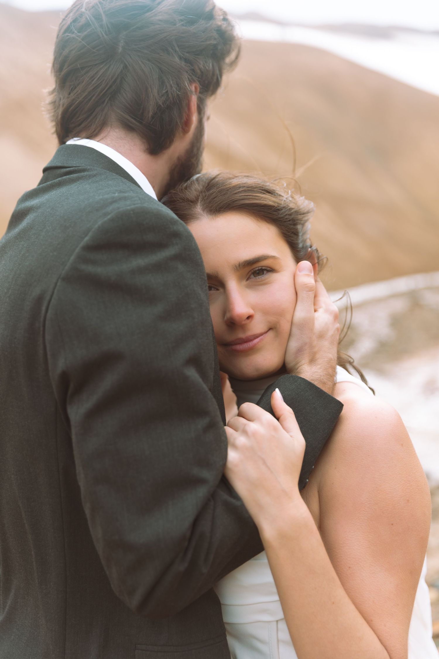 Bride rests her head on groom's chest as he cradles her face, with sandy mountains in the background.