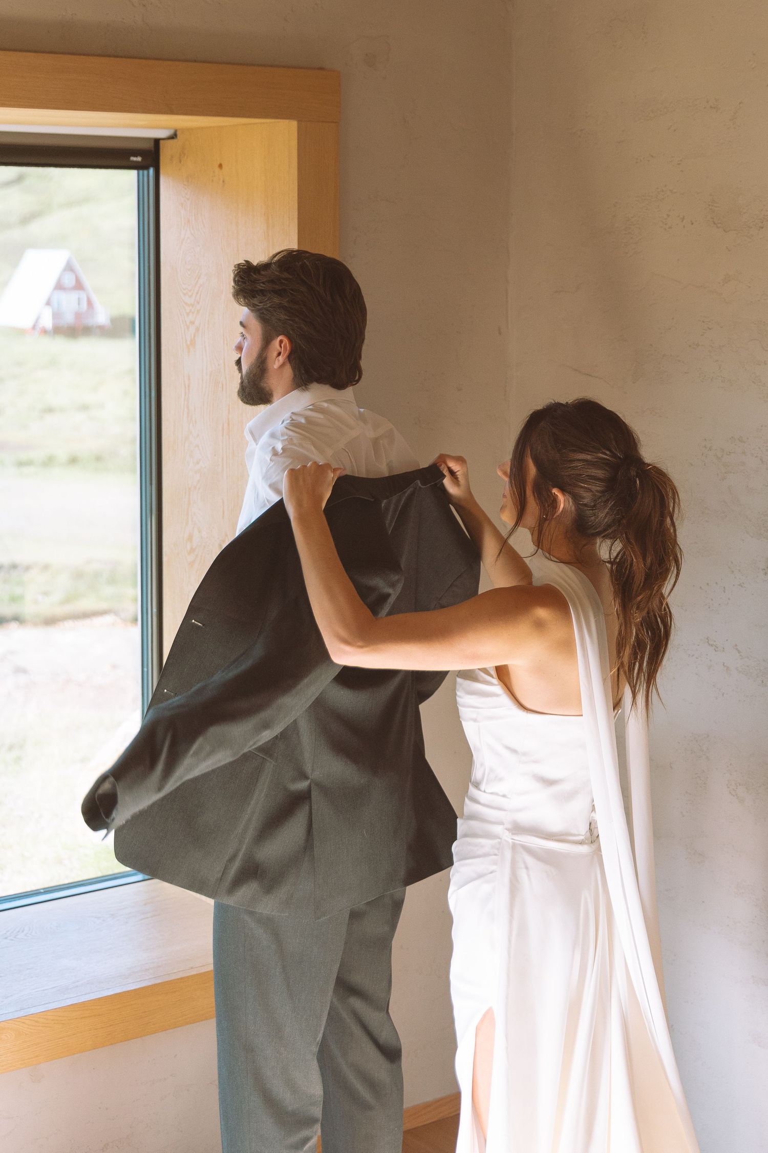 Bride assisting groom with his suit jacket near a bright window with scenic views.