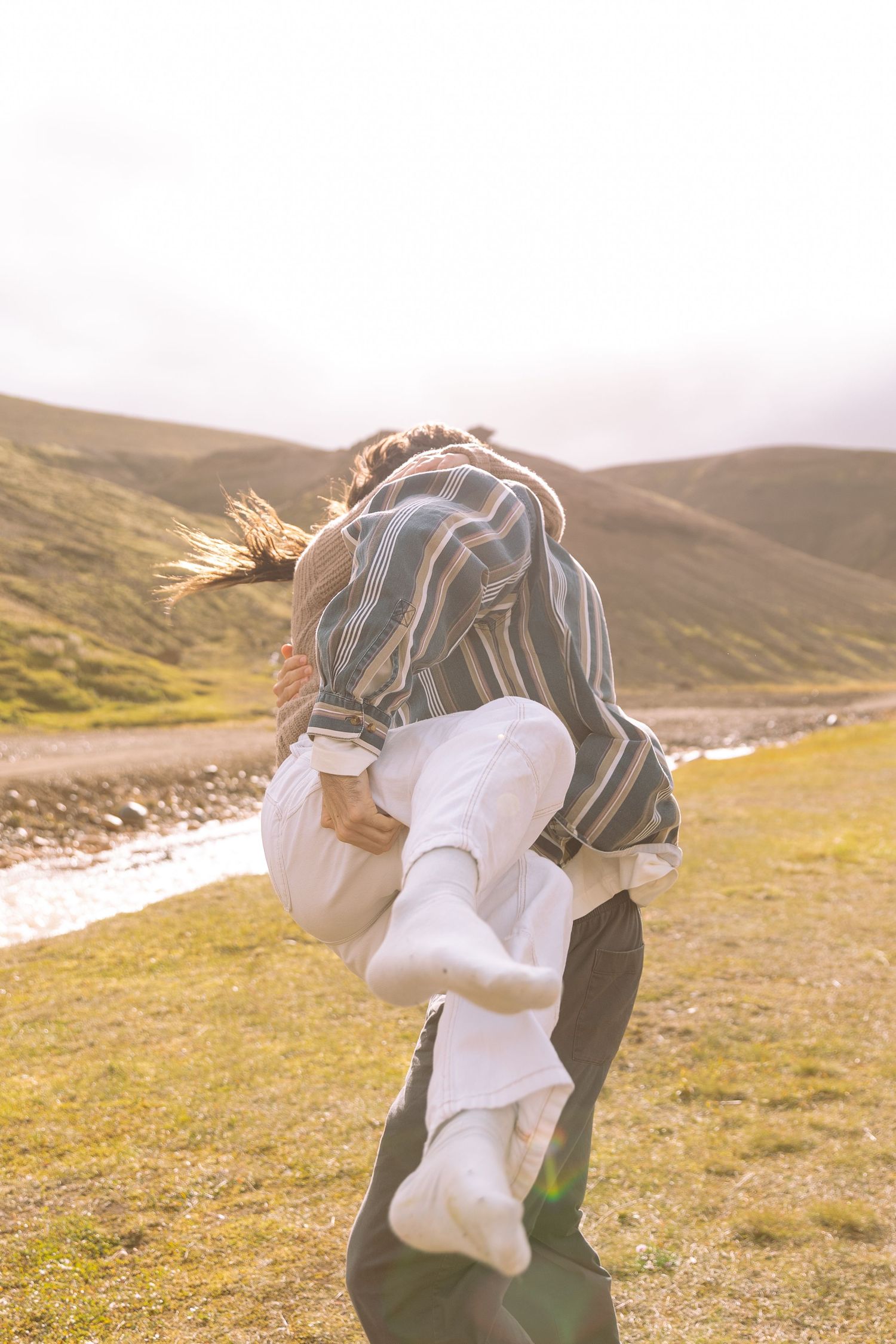 Couple embracing playfully as man lifts woman in golden sunlight beside a stream in Iceland.