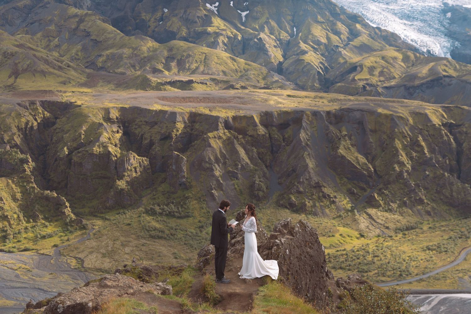 Wedding couple standing on rocky cliff edge with vast Icelandic highland canyon below.