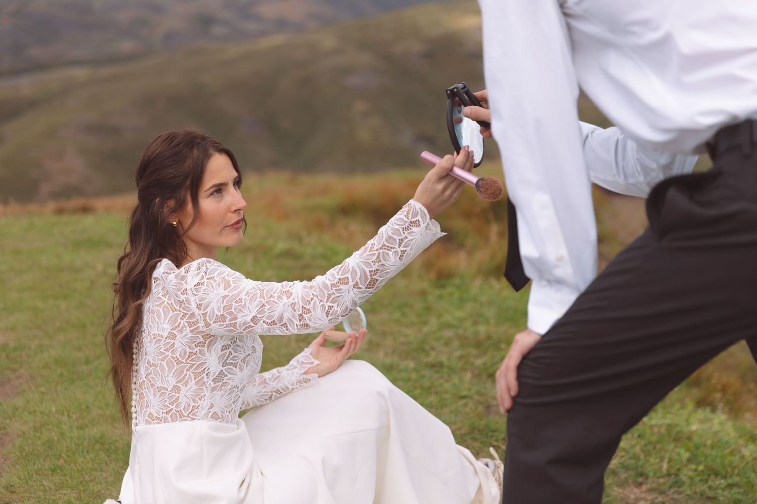 Bride in white lace gown reaches for camera held by groom during outdoor mountain wedding photoshoot.