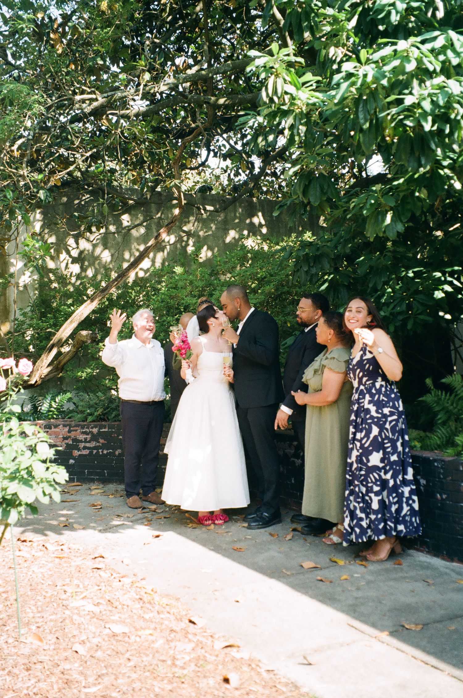 Bride and groom share a kiss surrounded by family at an outdoor garden wedding ceremony.