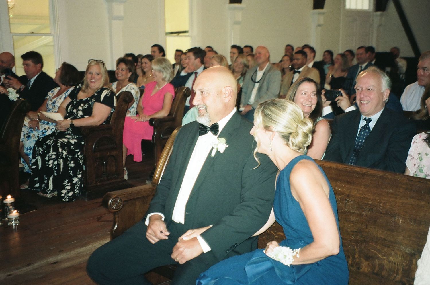 Guests seated in church pews during a wedding ceremony, with groom in tuxedo and bride in blue gown in foreground.