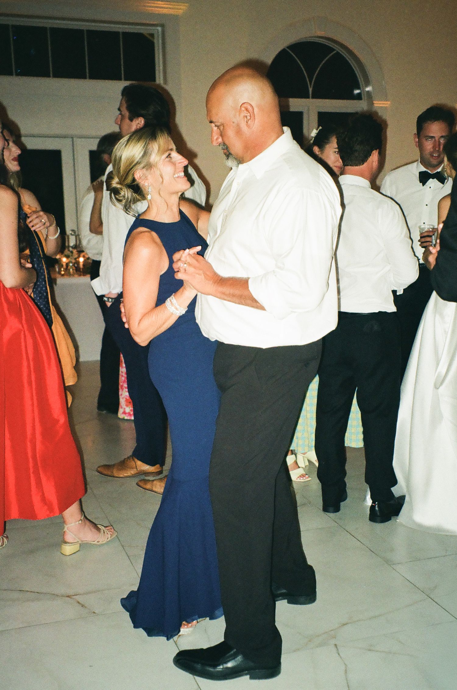 Smiling woman in navy gown dancing with man in white shirt at lively wedding reception.
