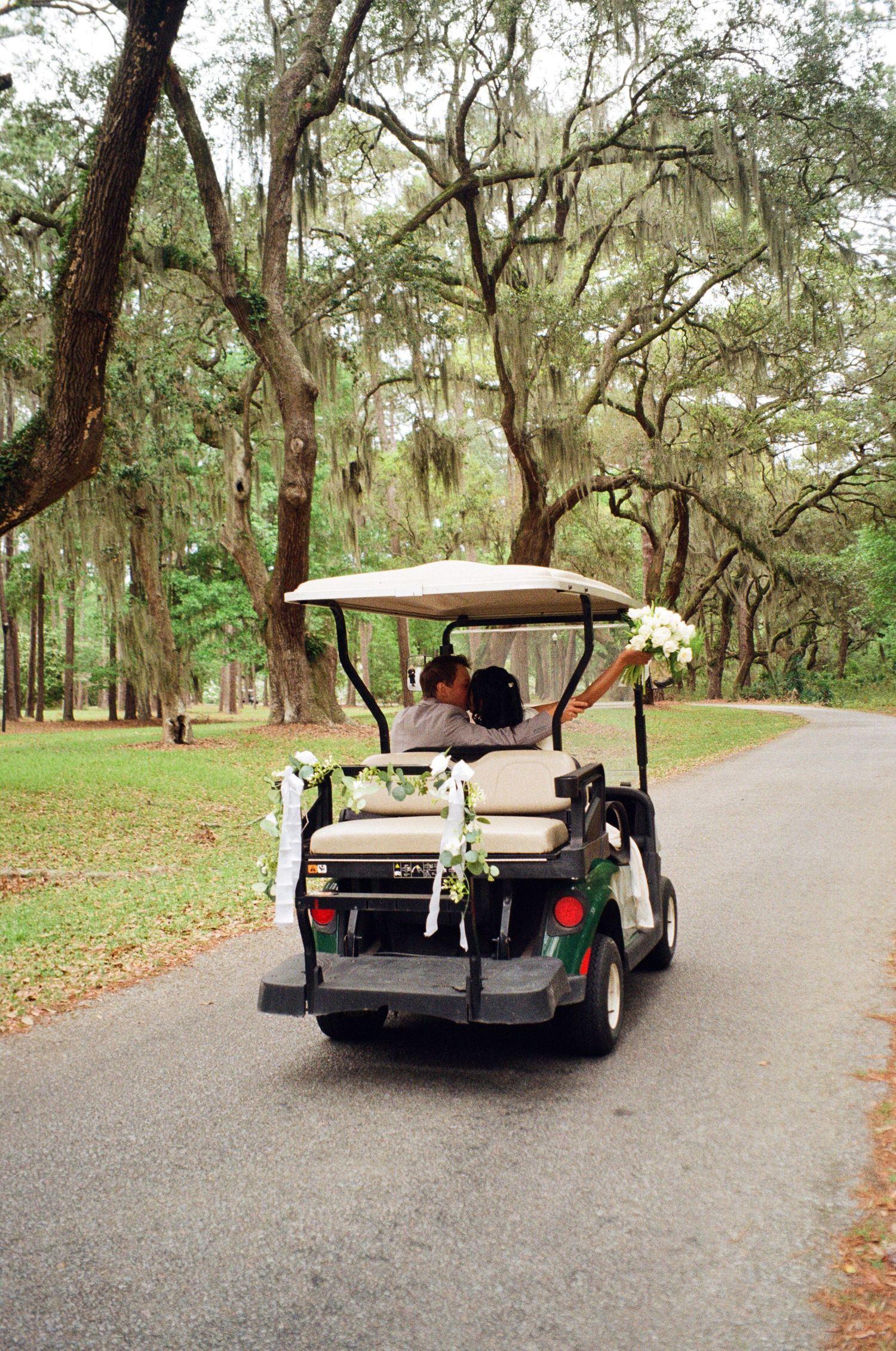 Decorated golf cart with white flowers drives down a tree-lined path at an outdoor wedding venue.