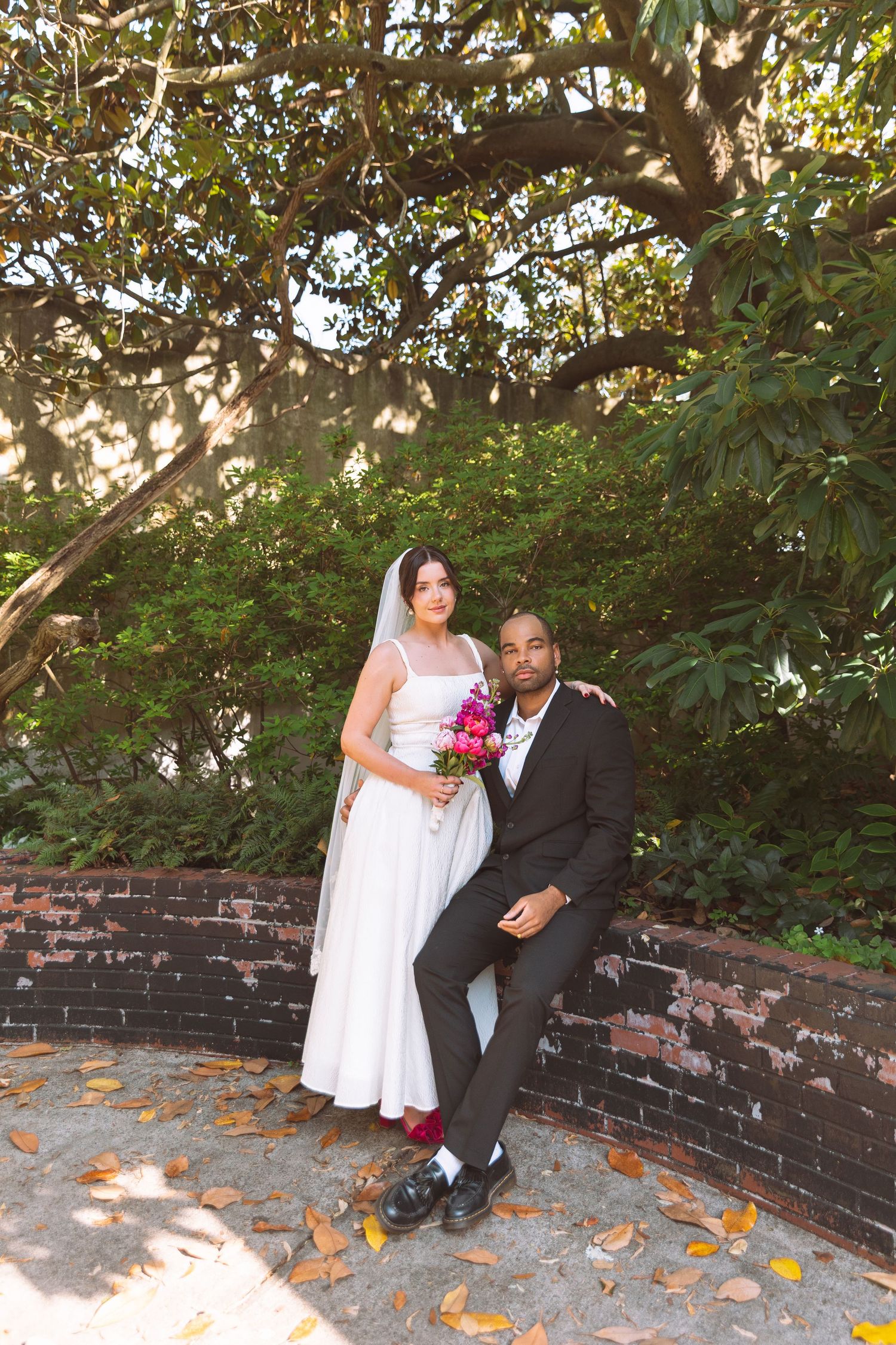 Interracial bride and groom posing under a magnolia tree with a pink bouquet at their wedding.