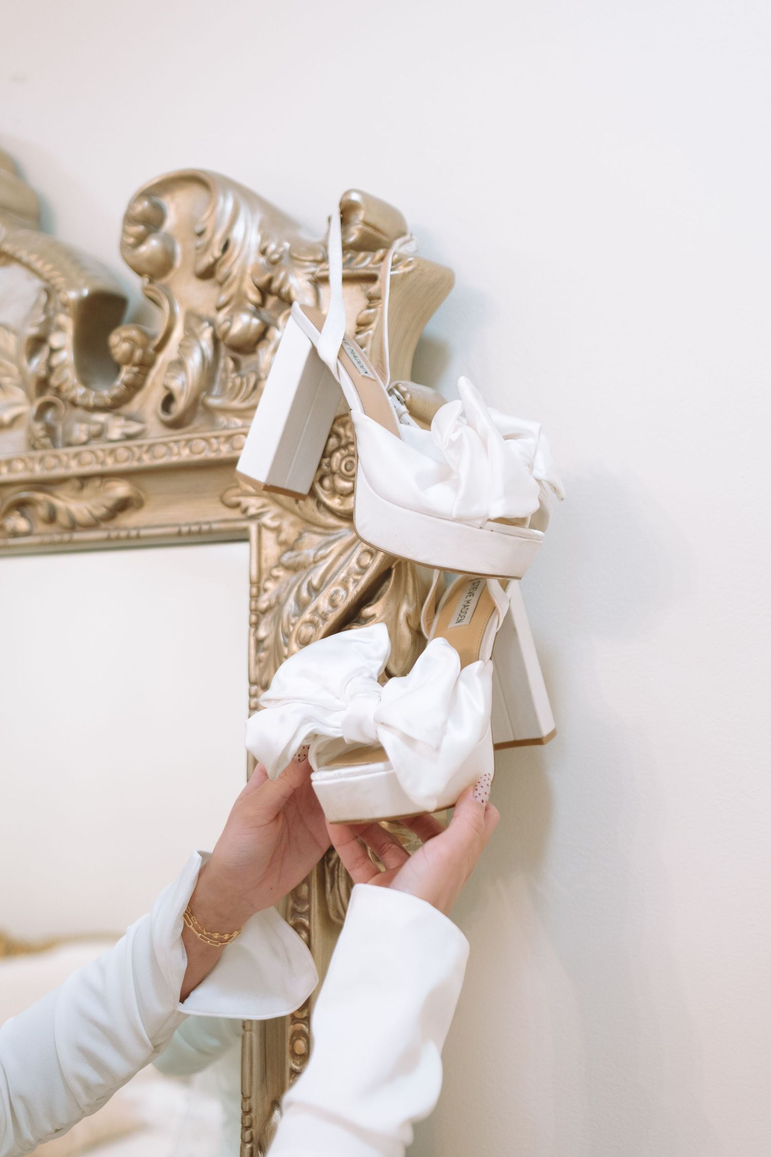 Bride holds white bow-adorned heels against an ornate gold-framed mirror.
