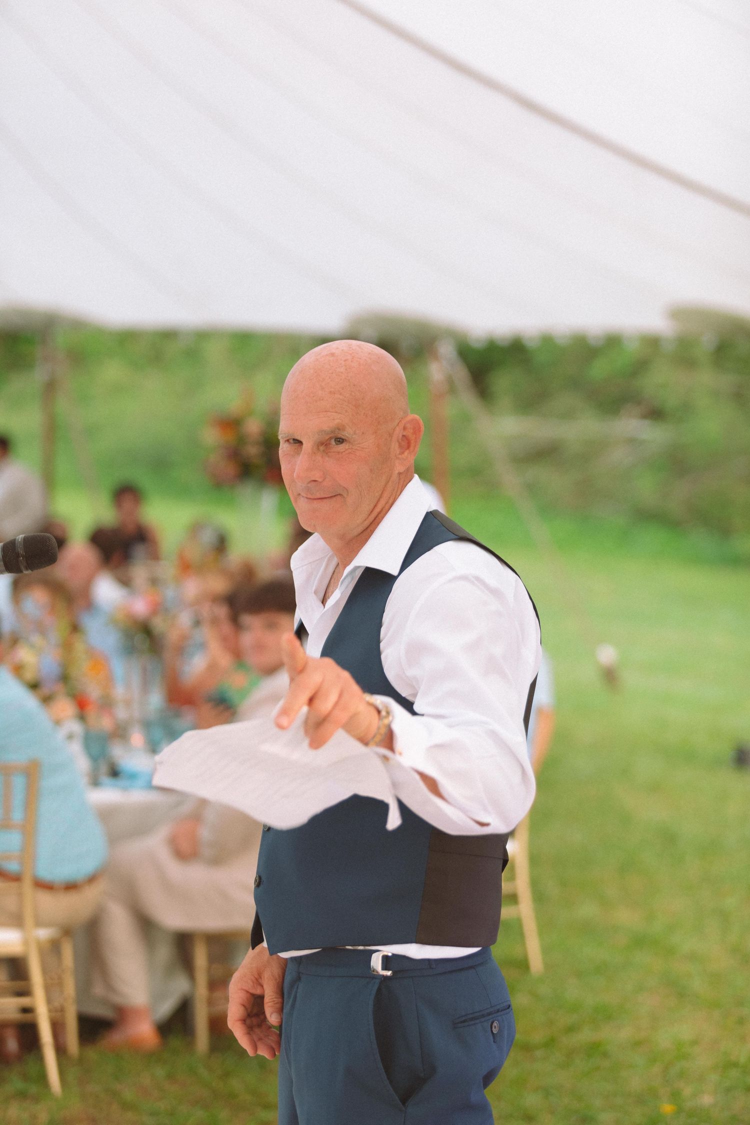Bald man in navy vest delivers speech holding papers at outdoor wedding reception under white tent.