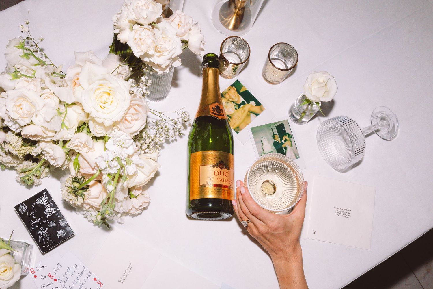 Hand holding a champagne flute beside a Veuve Clicquot bottle surrounded by white roses and photos.