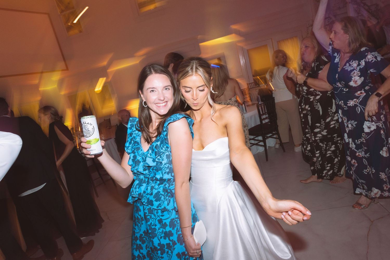 Bride in white gown dancing with guest in blue floral dress at wedding reception.