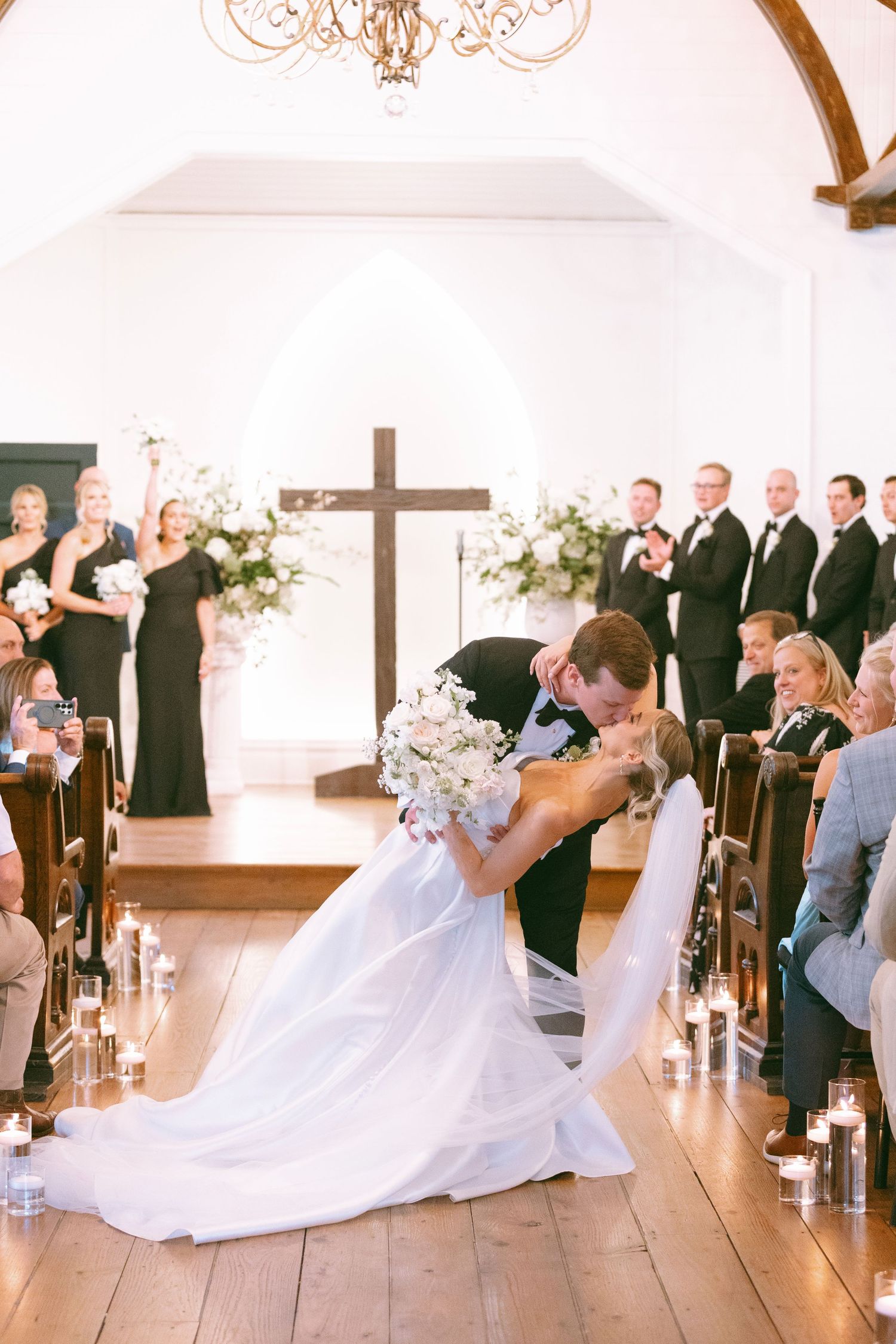 Bride and groom share a dip kiss at the altar in a white chapel with cross backdrop.