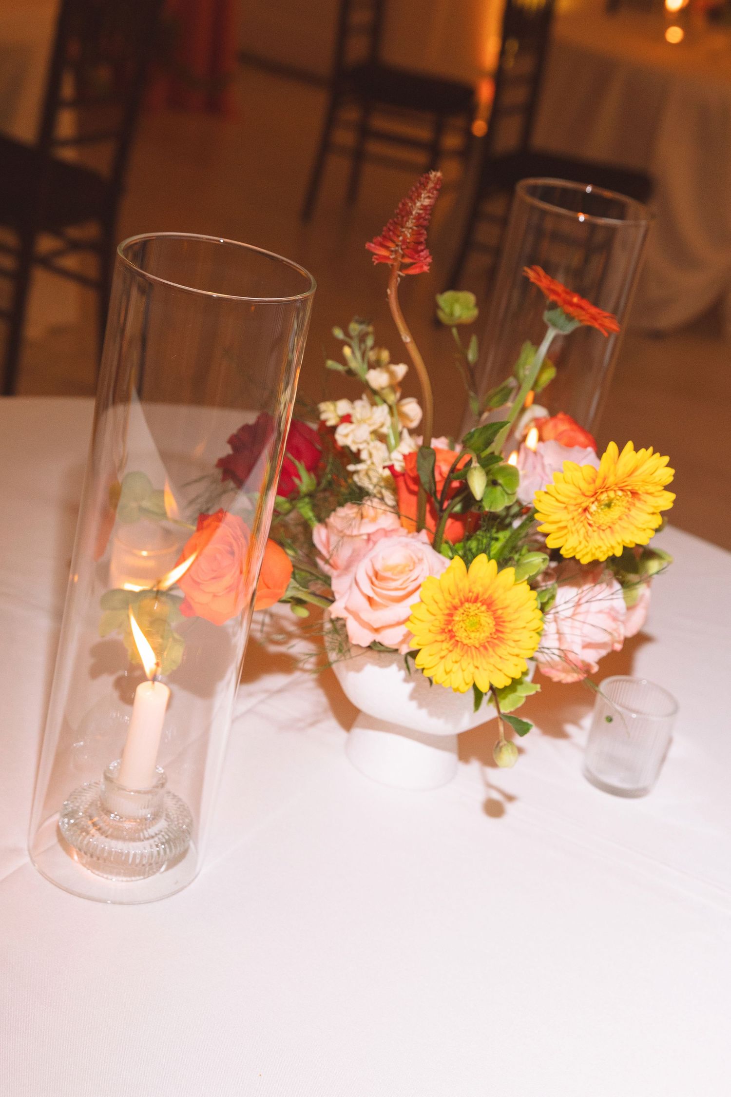 Elegant wedding centerpiece with yellow and pink flowers, candle in glass hurricane vase on white tablecloth.