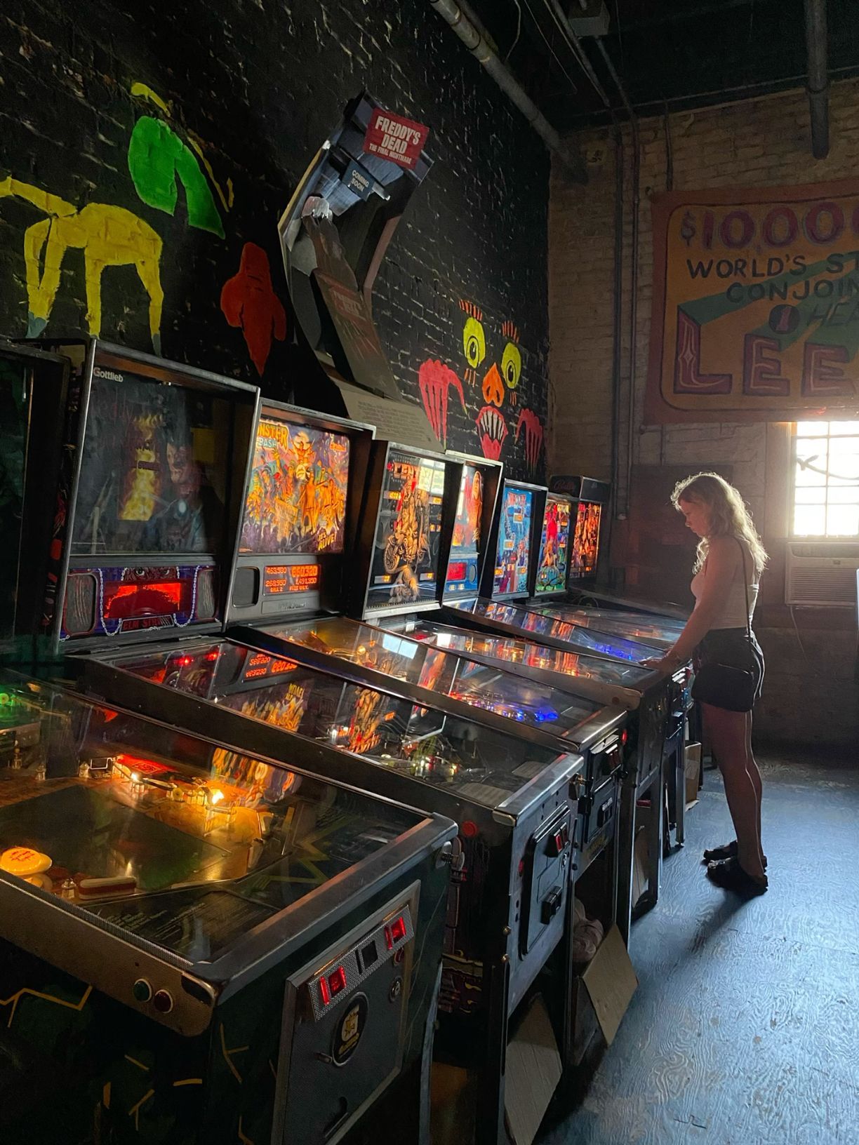 Woman playing vintage pinball machines in a dimly lit arcade bar with colorful neon artwork.