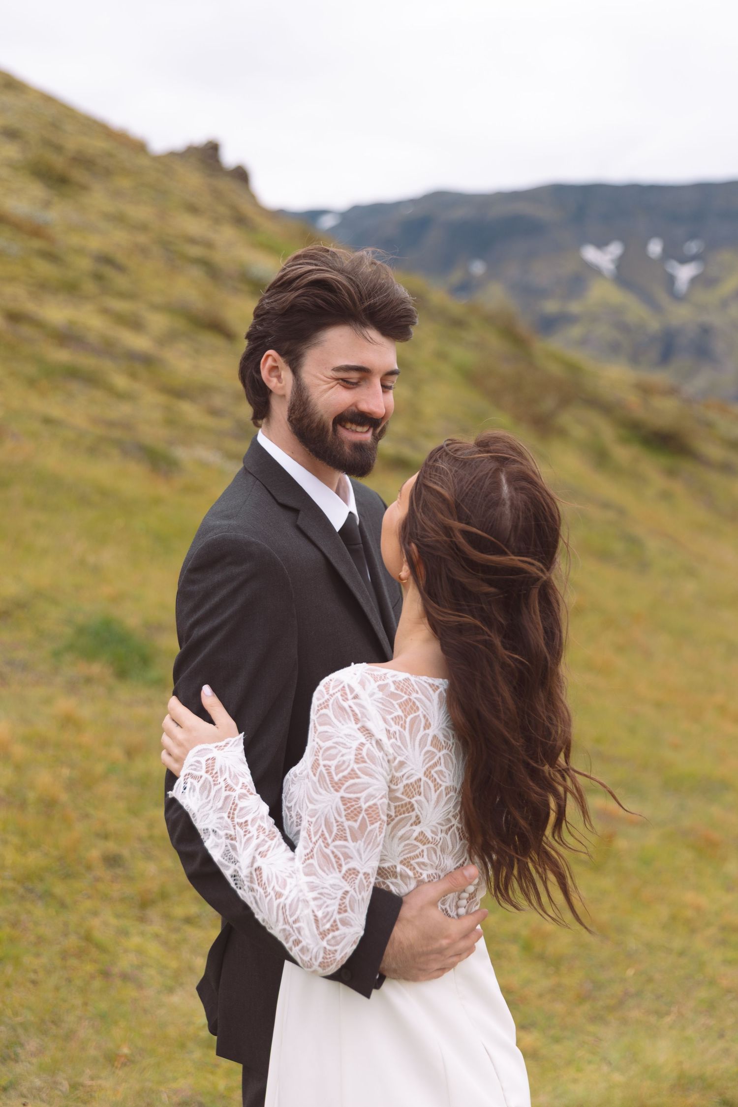 Smiling bearded groom in dark suit embraces bride in lace dress on green hillside.