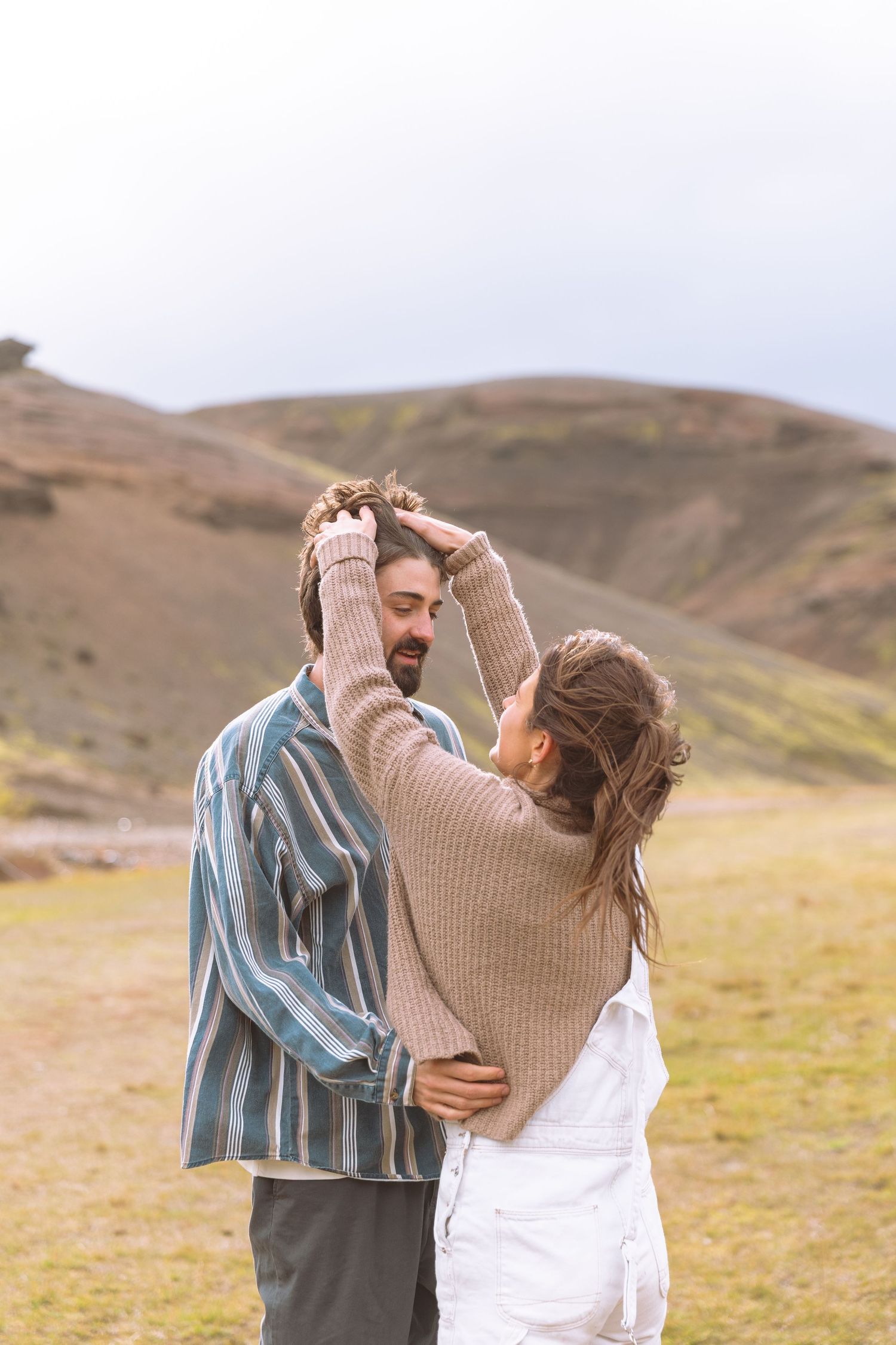 Woman playfully fixing bearded mans hair while standing together in scenic green hills.